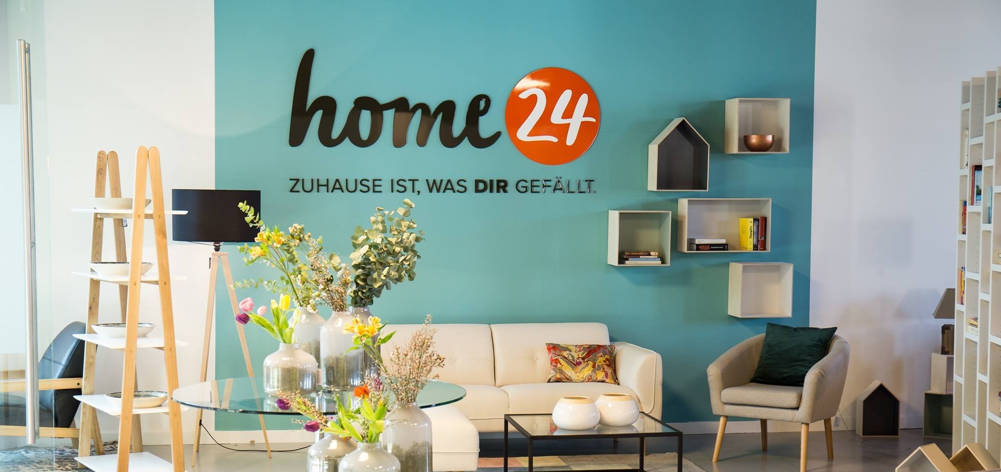 The home 24 logo is printed on the wall of a well-decorated room with lovely vasts, flowers, and a sofa, among other things.