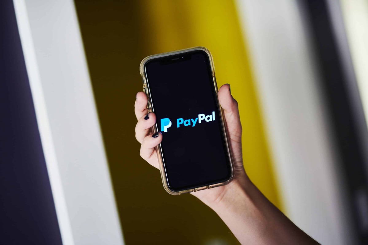 A girl with black nail polish holds a cellphone with the PayPal logo on a black background.