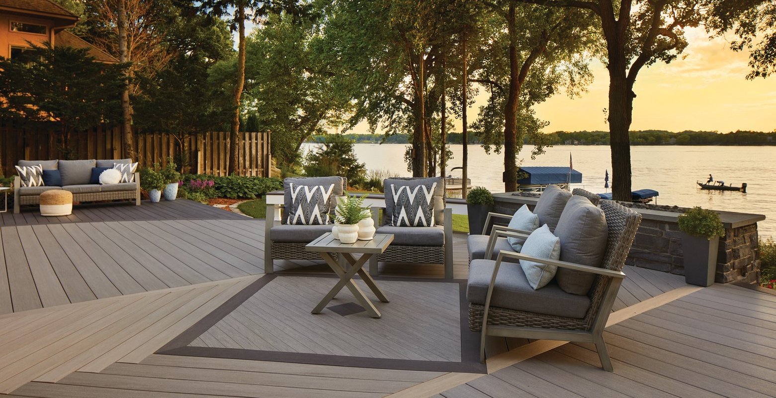 An outdoor deck overlooking a view of a lake surrounded by trees, is tiled with brown and grey woodwork, along with matching patio furniture during the afternoon