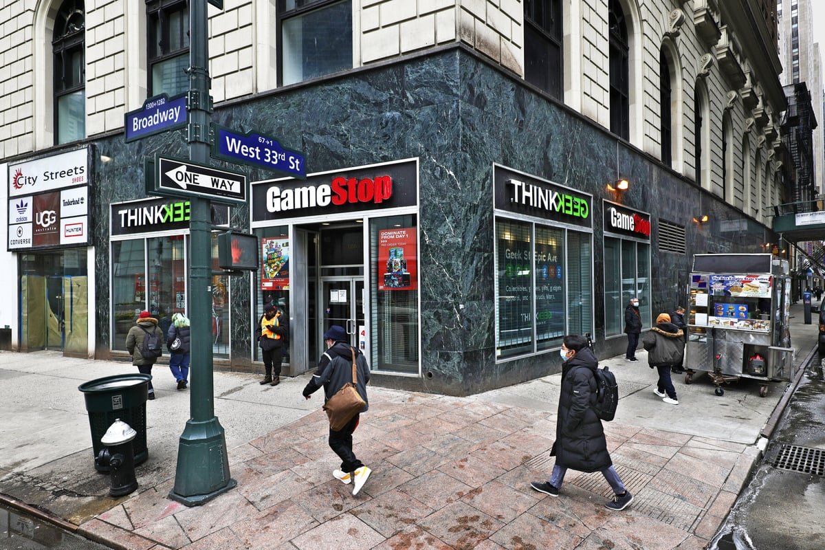 A stonewalled GameStop store at the corner of a busy street displays posters of multiple games on glass windows