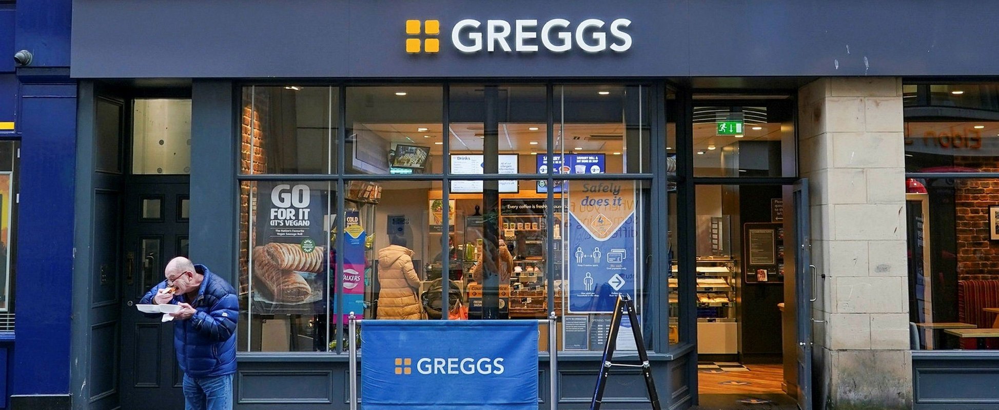 In front of the well-organized Greggs Bakery Store, a man is eating a Greggs' sandwich.
