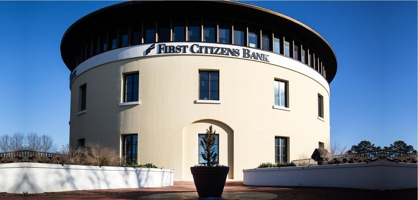 The image depicts the First Citizens Bank building, that has off-white walls and blue mirror windows.