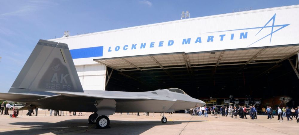 A fighter plane is parked in front of an airport apron with Lockheed Martin's logo on the top with crowds of people looking on